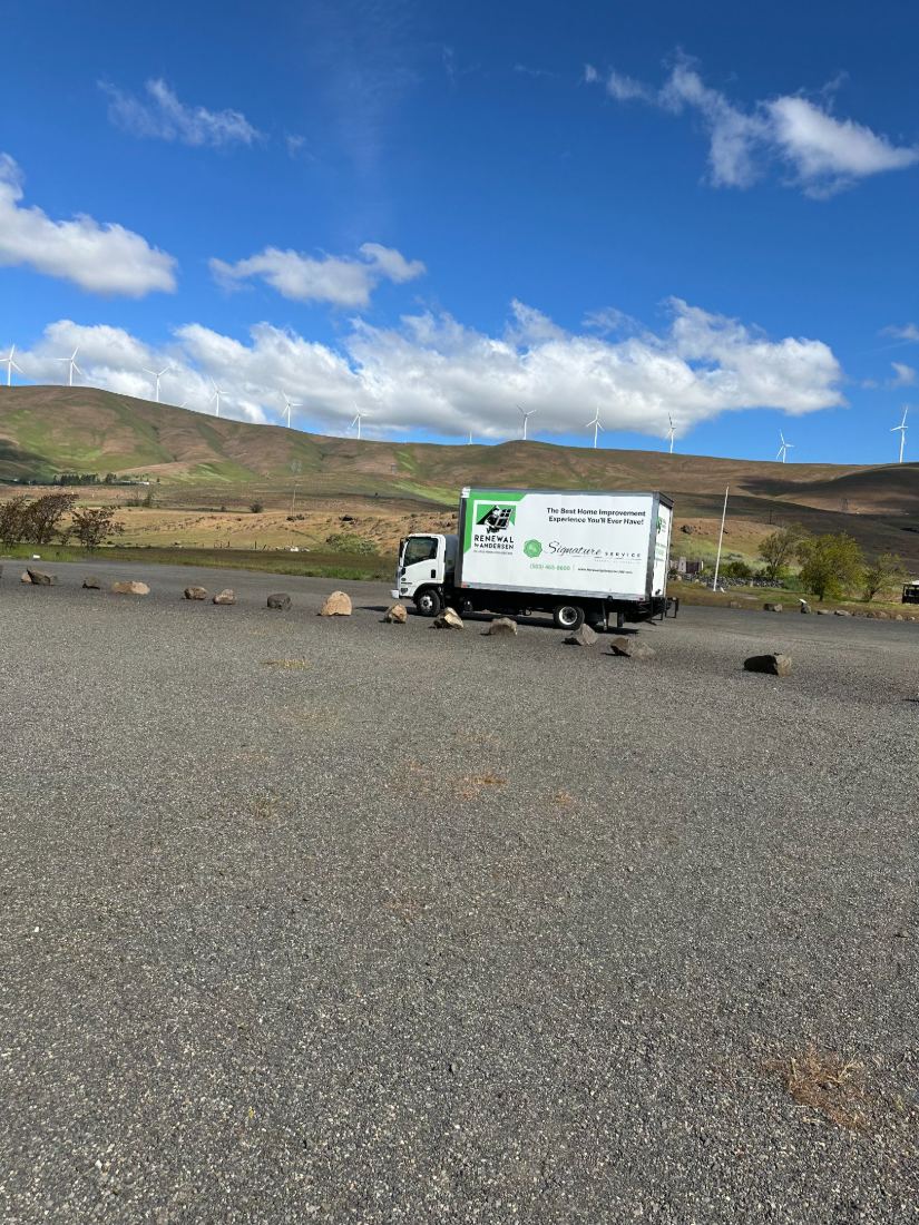 A Renewal by Andersen delivery truck parked on a gravel lot below wind-turbine hills and open sky